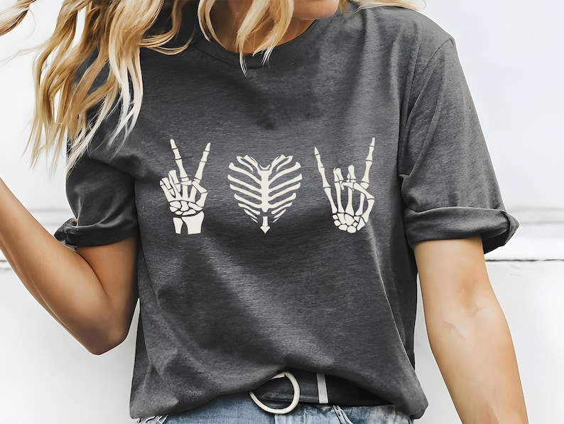 May include: Dark gray t-shirt with a white graphic design featuring a peace sign hand, a rib cage heart, and a rock-on hand gesture. The shirt has short sleeves and a crew neck.