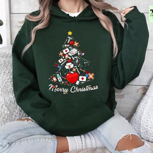 May include: Dark green hoodie with a Christmas tree design made of medical supplies, including a red heart, bandages, and syringes. The words "Merry Christmas" are written in white script below the tree. A yellow star tops the tree.