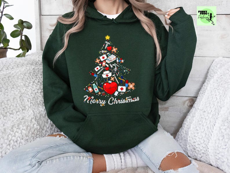 May include: Dark green hoodie with a Christmas tree design made of medical supplies, including a red heart, bandages, and syringes. The words "Merry Christmas" are written in white script below the tree. A yellow star tops the tree.