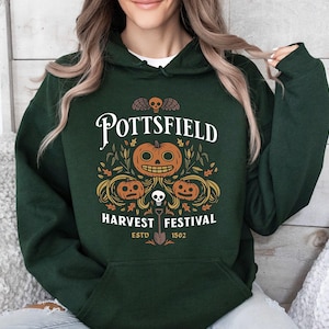 May include: A dark green hooded sweatshirt featuring a graphic print with the text POTTSFIELD HARVEST FESTIVAL ESTD 1502. The design includes three orange jack-o'-lanterns, a small skull, a shovel, and autumn leaves in white and orange.
