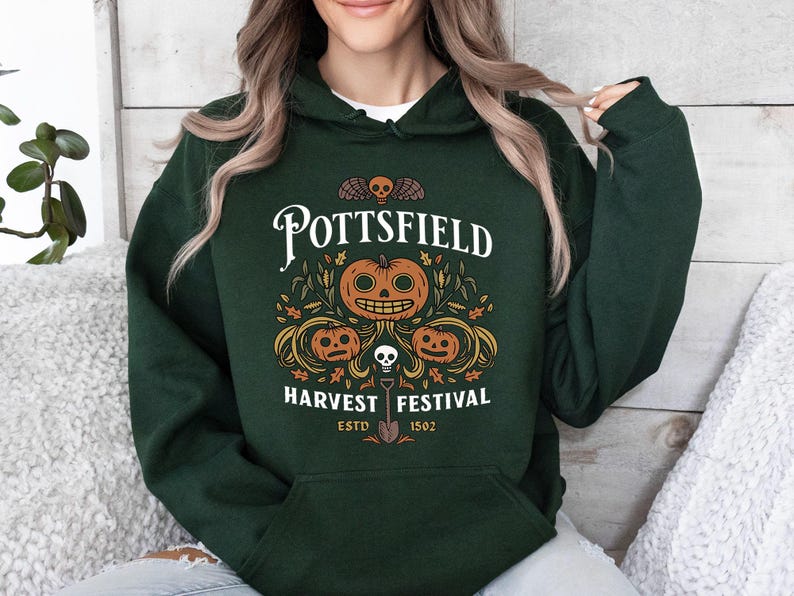 May include: A dark green hooded sweatshirt featuring a graphic print with the text POTTSFIELD HARVEST FESTIVAL ESTD 1502. The design includes three orange jack-o'-lanterns, a small skull, a shovel, and autumn leaves in white and orange.