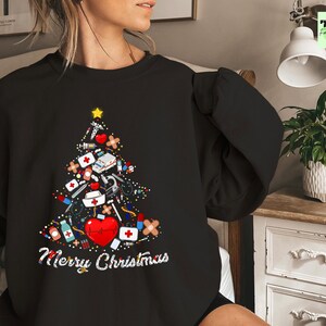 May include: Black sweatshirt featuring a Christmas tree design made of medical supplies, a red heart, and a gold star. The words "Merry Christmas" are written in white script below the tree. A festive holiday sweater.