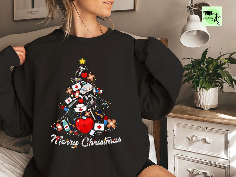 May include: Black sweatshirt featuring a Christmas tree design made of medical supplies, a red heart, and a gold star. The words "Merry Christmas" are written in white script below the tree. A festive holiday sweater.