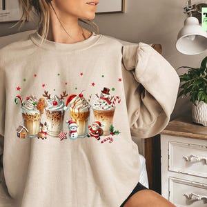 May include: Beige sweatshirt featuring a festive graphic of Christmas-themed coffee drinks. Each drink is adorned with whipped cream, candy canes, and holiday characters like gingerbread men, snowmen, and Santa Claus. Red and green stars and dots are scattered around the drinks.