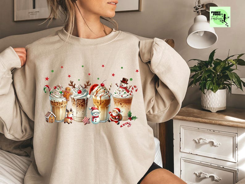 May include: Beige sweatshirt featuring a festive graphic of Christmas-themed coffee drinks. Each drink is adorned with whipped cream, candy canes, and holiday characters like gingerbread men, snowmen, and Santa Claus. Red and green stars and dots are scattered around the drinks.