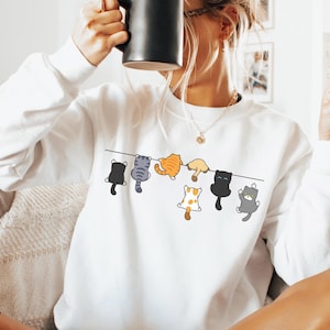 Pode incluir: Camisola branca com um design l&uacute;dico de gatos de desenho animado pendurados num varal. Os gatos s&atilde;o de v&aacute;rias cores, incluindo preto, cinza, laranja e branco, adicionando um toque divertido &agrave; roupa casual.
