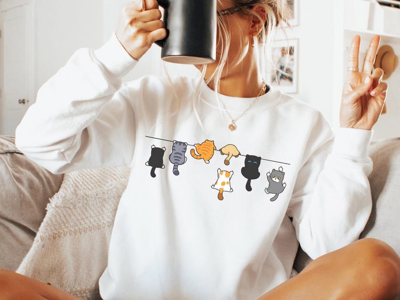 Pode incluir: Camisola branca com um design l&uacute;dico de gatos de desenho animado pendurados num varal. Os gatos s&atilde;o de v&aacute;rias cores, incluindo preto, cinza, laranja e branco, adicionando um toque divertido &agrave; roupa casual.