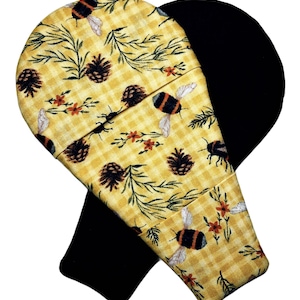 May include: A fabric oven mitt featuring a yellow and white gingham pattern with a print of bumblebees, pinecones, and small orange flowers. The mitt has a rounded top and a solid black fabric lining visible on the interior and the back side.