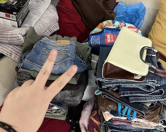 May include: A pile of clothing, including jeans, shirts, and a handbag. The jeans are various shades of blue, and the handbag is cream-colored with a brown base and handle. Other items include a video game box and a blue plastic bag.