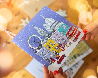 May include: A blue Christmas card with a snowy London skyline illustration. The card features iconic landmarks like Big Ben, the London Eye, and a red double-decker bus.