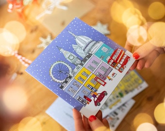 May include: A blue Christmas card with a snowy London skyline illustration. The card features iconic landmarks like Big Ben, the London Eye, and a red double-decker bus.