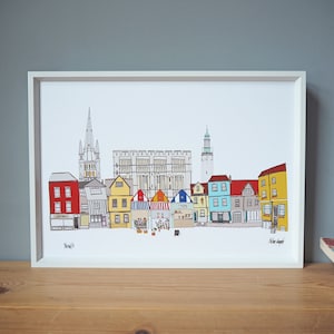 以下が含まれることがあります： 教会、城、時計塔のある街の水平線を表すカラフルなイラスト。建物は黄色、赤、青、緑の様々な色合いで描かれています。イラストは白い額縁に入っています。