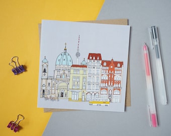 May include: A white card with a hand-drawn illustration of a cityscape in Berlin, Germany. The illustration features a church with a blue dome, a tall tower, and several buildings with red roofs. The card is on a brown envelope and a yellow surface.
