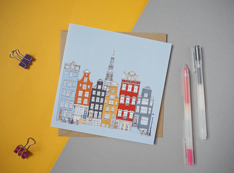 May include: A blue and white card with a hand-drawn illustration of colorful buildings in a city. The buildings are in a row and have different styles and colors. There is a church steeple in the center of the illustration.