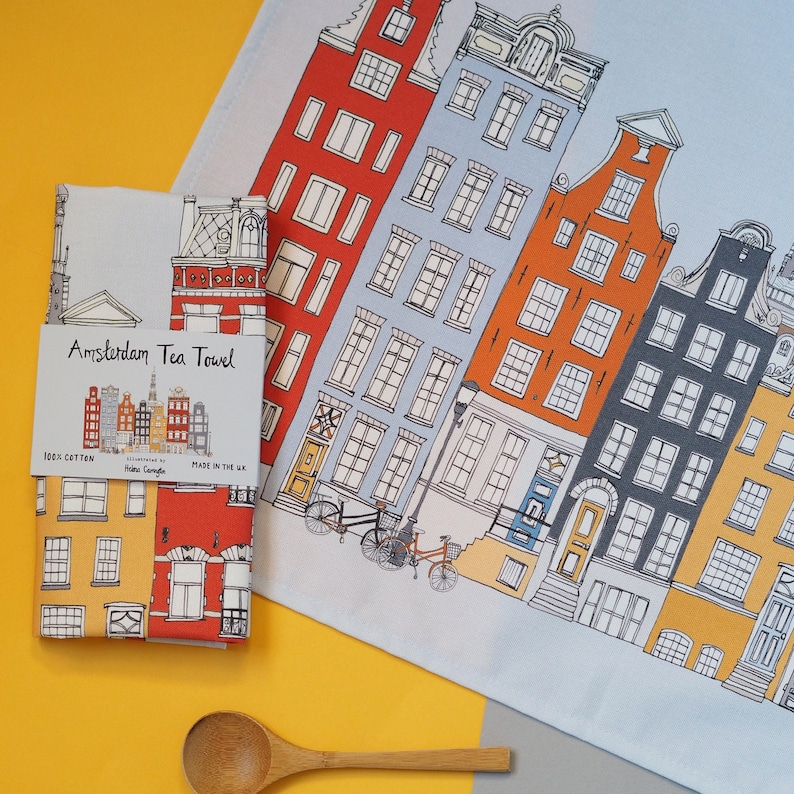 Pu&ograve; includere: Un asciugamano da t&egrave; blu e bianco con un'illustrazione colorata di edifici di Amsterdam. L'asciugamano &egrave; realizzato in 100% cotone e presenta il testo "Amsterdam Tea Towel" su uno stendardo bianco.