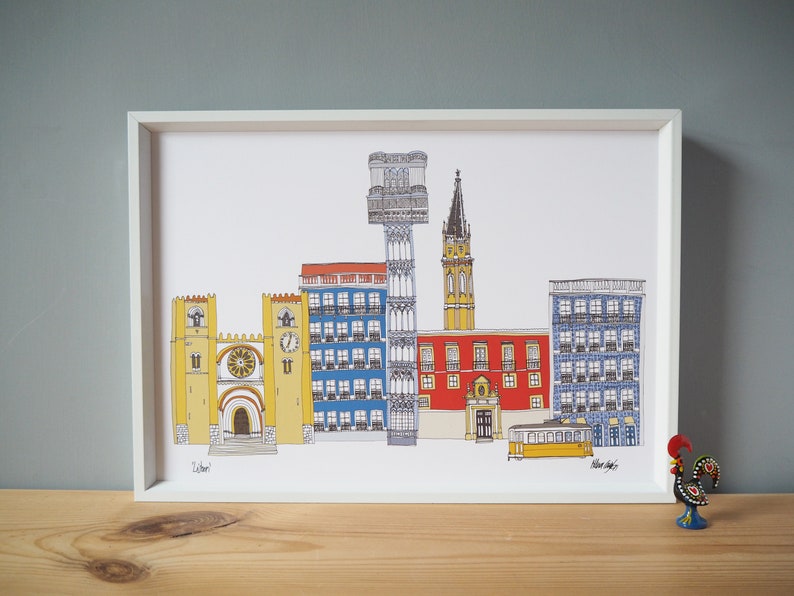 May include: A framed illustration of a cityscape with colorful buildings, including a cathedral, a tower, and a tram. The illustration is in a white frame and is on a wooden surface.