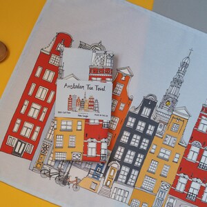 Pu&ograve; includere: Un asciugamano da t&egrave; bianco con un'illustrazione colorata di edifici di Amsterdam. L'asciugamano ha la scritta "Amsterdam Tea Towel" stampata su di esso.