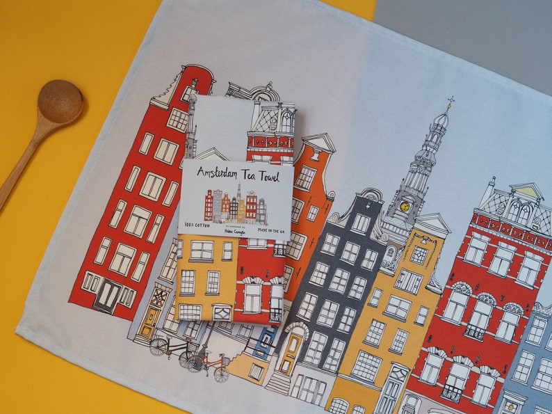 Pu&ograve; includere: Un asciugamano da t&egrave; bianco con un'illustrazione colorata di edifici di Amsterdam. L'asciugamano ha la scritta "Amsterdam Tea Towel" stampata su di esso.