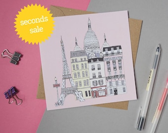 May include: A pink card with a black and white illustration of Parisian landmarks, including the Eiffel Tower, Sacré-Coeur Basilica, and a cafe. The card is surrounded by a brown envelope and two pens.