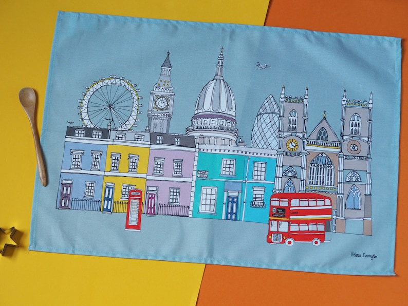 May include: A blue tea towel with a colorful illustration of London landmarks, including Big Ben, the London Eye, St. Paul's Cathedral, and a red double-decker bus. The text "Helena Carrington" is printed in the bottom right corner.