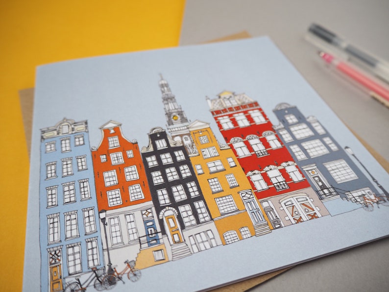 May include: A colorful illustration of a row of buildings with a blue sky background. The buildings are drawn in a cartoon style and have different colors, including red, yellow, blue, and black. There are bicycles in front of the buildings.