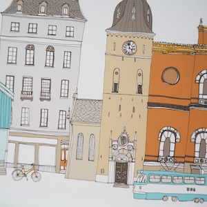 Pu&ograve; includere: Un'illustrazione colorata di una strada cittadina con edifici di vari stili, tra cui una chiesa con un campanile, un edificio con una vetrina e un edificio con un balcone. Un tram blu e bianco &egrave; parcheggiato di fronte agli edifici.
