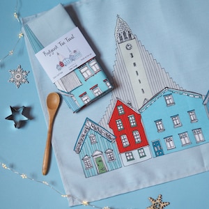 May include: A blue tea towel featuring a white illustration of Hallgrímskirkja church and other buildings in Reykjavik, Iceland. The text "Reykjavik Tea Towel" is printed on the towel.