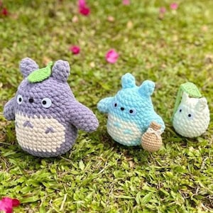 May include: Three crocheted figures of a popular animated character are displayed on a bed of green grass. The figures are in shades of purple, blue, and white, with black eyes and leaf accents. One figure holds a small brown bag.