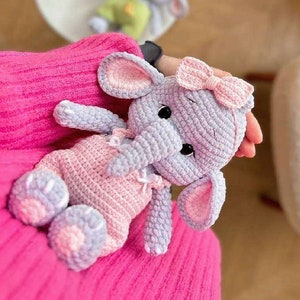 以下が含まれることがあります： ピンクのドレスとピンクのリボンをつけた、かぎ針編みのかわいいぬいぐるみゾウ。ゾウはグレーで、ピンクの耳と足をしています。