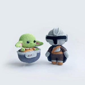 May include: A green and brown crocheted plush toy of a character from the Star Wars franchise, sitting in a grey crocheted spaceship.  The character is wearing a brown and orange outfit.  The spaceship is floating in the air.  The character is looking to the right.  The spaceship is facing the left.  The background is white.
