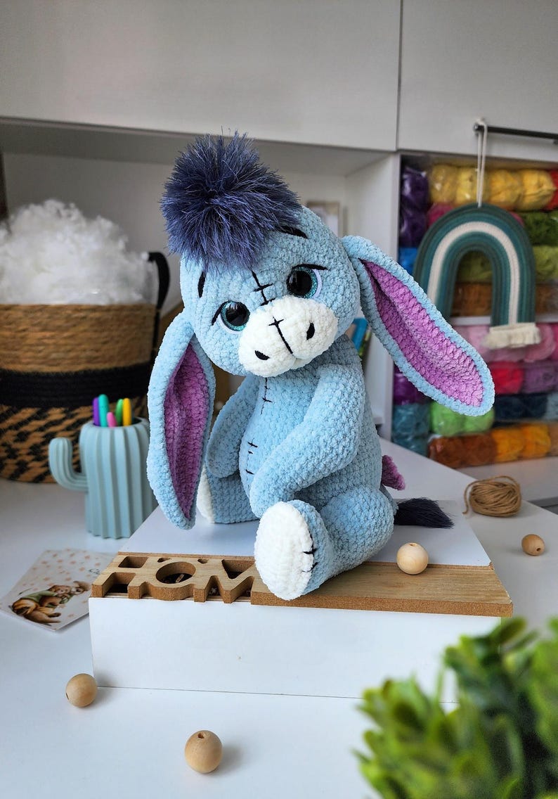 May include: A handmade plush toy of a blue donkey with a purple mane and ears. The stuffed animal has large, expressive eyes and is sitting on a white surface. The toy is a character from a popular children's story.
