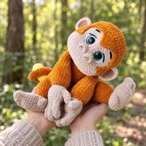 May include: A plush monkey toy crafted from soft orange and beige yarn. The monkey features large, round, dark eyes with bright blue irises, a rounded face, and small ears. It has a textured, fuzzy appearance with a small yellow tail visible.