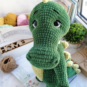 Pode incluir: Um brinquedo de dinossauro verde de crochê feito à mão com uma expressão amigável. O bicho de pelúcia tem uma superfície texturizada, detalhes amarelos e olhos grandes e expressivos. Está sentado em um livro com ilustrações florais, sugerindo um tema infantil.