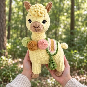 May include: A handmade crochet llama plush toy in pale yellow with a brown muzzle and inner ears. It features three yarn pom-poms in sage green, tan, and dusty pink around the neck, and a decorative saddle blanket with a tassel in green and pink.