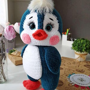 May include: A handmade, plush penguin doll with a blue and white body, red feet, and a white fluffy head. The penguin has large, expressive eyes with long eyelashes, pink cheeks, and a red beak. It is a cute, whimsical toy.