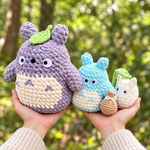 May include: Three crocheted plush figures with rounded ears and white bellies. The largest is purple with a green leaf on its head. The medium figure is light blue, and the smallest is white with a green leaf and a small tan drawstring bag.