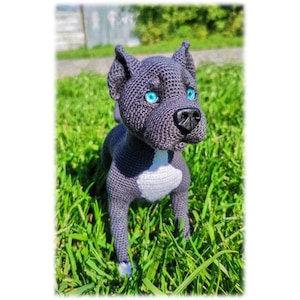 May include: A crocheted grey and white pit bull dog toy with blue eyes. The toy is sitting on a bed of green grass.