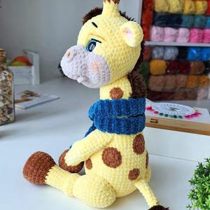 May include: A crocheted giraffe plush toy made of soft yellow yarn with brown spots, a brown mane, and brown hooves. The giraffe has a light gray muzzle, blue eyes, and is wearing a textured, dark blue crocheted scarf around its neck.