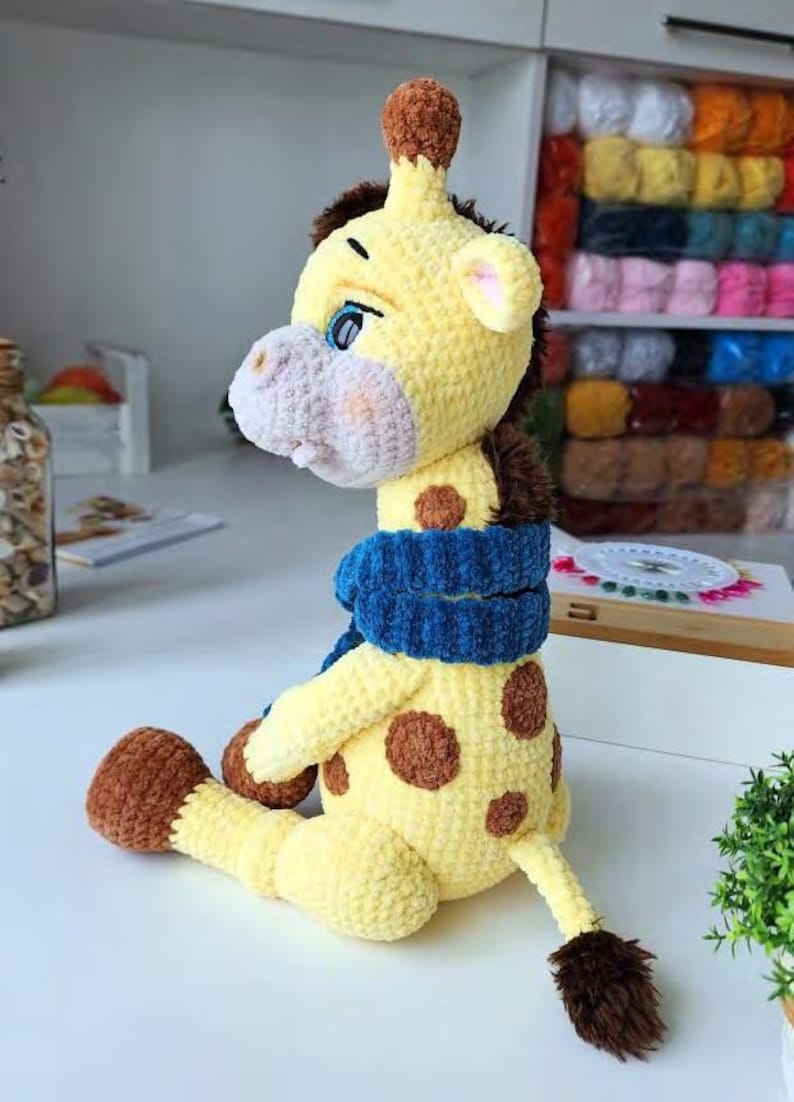 May include: A crocheted giraffe plush toy made of soft yellow yarn with brown spots, a brown mane, and brown hooves. The giraffe has a light gray muzzle, blue eyes, and is wearing a textured, dark blue crocheted scarf around its neck.