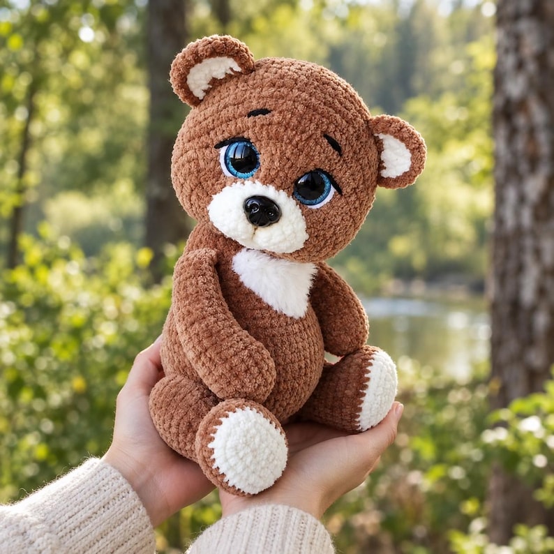 May include: A brown crocheted teddy bear with large blue eyes, a black nose, and white accents on the ears, chest, and feet. The stuffed toy has a soft, textured yarn finish and is crafted with a rounded head and body in a seated position.
