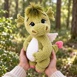 May include: A crocheted dragon plush toy featuring a light green textured body, a white belly, and small white wings. The creature has large dark eyes, a tuft of olive green hair on its head, and a small pink heart-shaped detail on its tail tip.