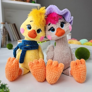 Può includere: Due bambole di uccelli fatte a mano all'uncinetto. Una è gialla con una sciarpa blu e capelli gialli, l'altra è beige con un copricapo viola e rosa. Entrambe hanno piedi e becchi arancioni e occhi blu. Sono sedute su una superficie bianca.