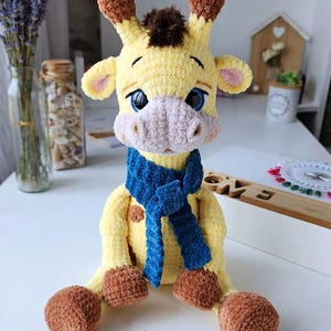 May include: A crocheted giraffe plush toy with yellow fur, brown spots, and a dark brown tuft of hair on its head. The giraffe has large blue eyes, a light pink muzzle, and brown hooves. It is wearing a textured, dark blue knitted scarf around its neck.