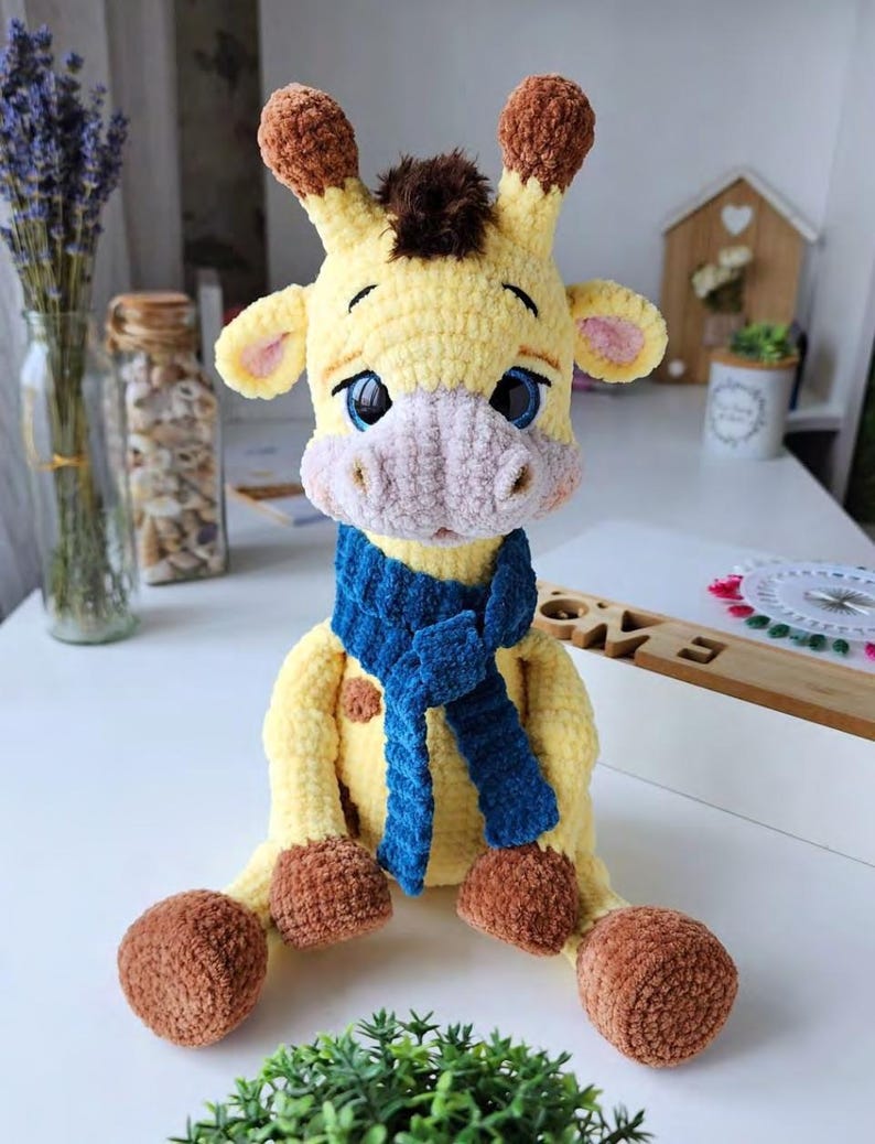May include: A crocheted giraffe plush toy with yellow fur, brown spots, and a dark brown tuft of hair on its head. The giraffe has large blue eyes, a light pink muzzle, and brown hooves. It is wearing a textured, dark blue knitted scarf around its neck.