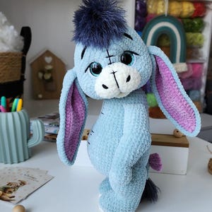 May include: A handmade plush toy of Eeyore, the donkey from Winnie the Pooh. The plush is light blue with a dark blue mane, pink inner ears, and a white muzzle. The toy is made of soft, textured material.