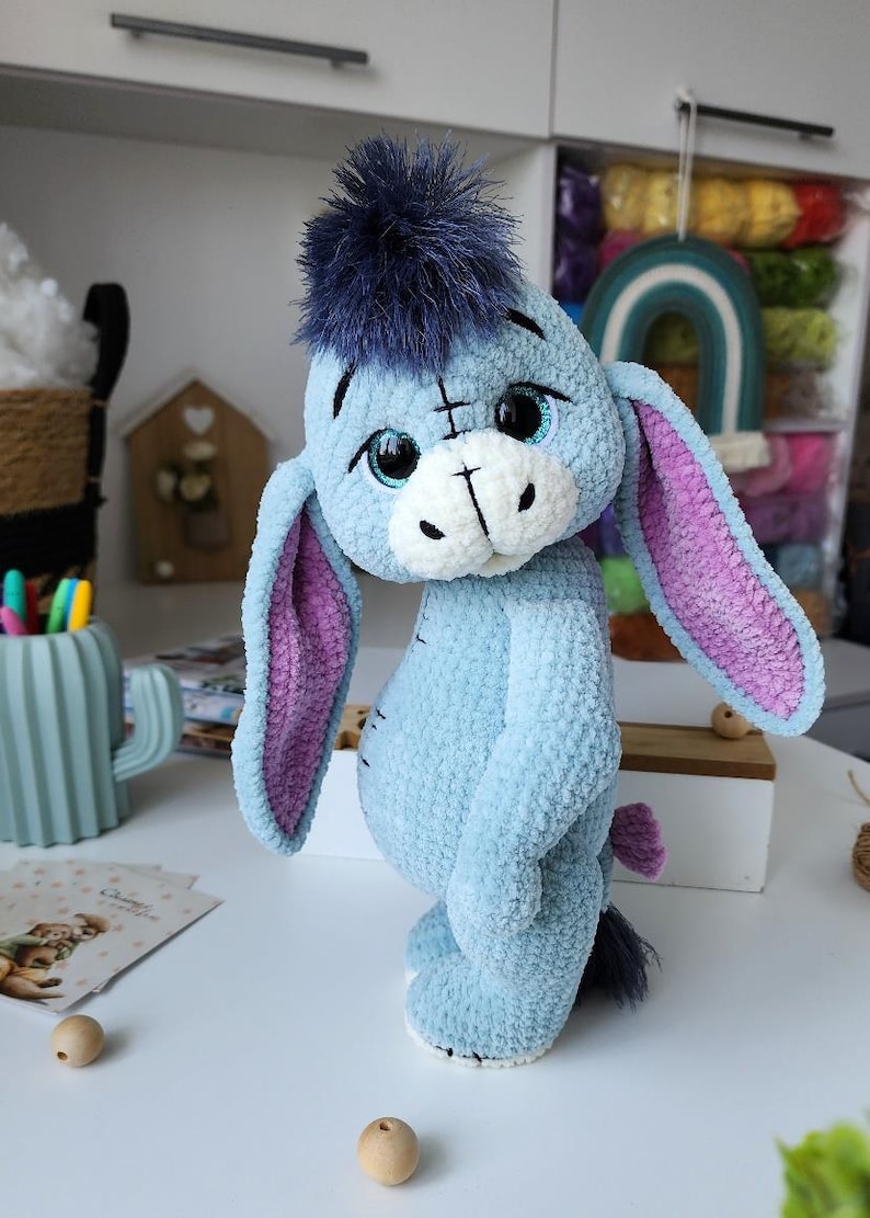 May include: A handmade plush toy of Eeyore, the donkey from Winnie the Pooh. The plush is light blue with a dark blue mane, pink inner ears, and a white muzzle. The toy is made of soft, textured material.