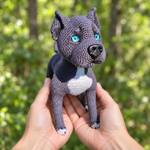 Puede incluir: Una figura de perro tejida a ganchillo en color gris oscuro con una mancha blanca en el pecho, patas con puntas blancas y ojos azul brillante. El peluche tiene una nariz de plástico negra y orejas puntiagudas, con textura de hilo.