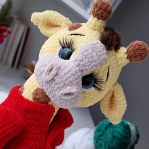 May include: A crocheted giraffe plush toy with yellow yarn, a light gray muzzle, and brown spots. The giraffe features large blue eyes with long black eyelashes, brown pom-pom horns, and a dark brown tuft of hair. It wears a bright red crocheted jacket.