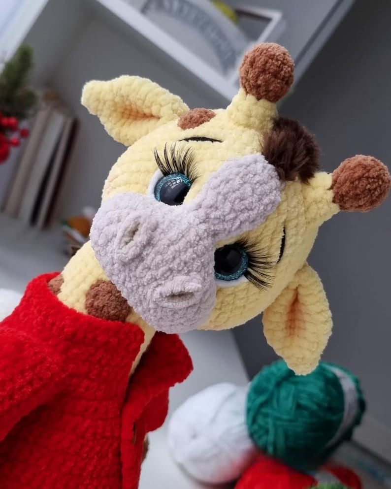 May include: A crocheted giraffe plush toy with yellow yarn, a light gray muzzle, and brown spots. The giraffe features large blue eyes with long black eyelashes, brown pom-pom horns, and a dark brown tuft of hair. It wears a bright red crocheted jacket.