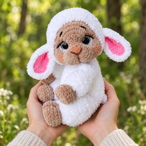 May include: A plush crochet lamb toy featuring a white fuzzy body, a light brown face, and large dark eyes. The lamb has long, floppy ears with bright pink inner accents, small brown paws, and a soft, textured yarn construction throughout.